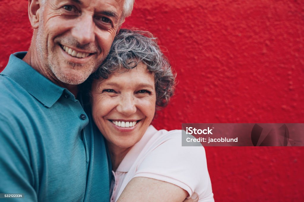 Cheerful mature couple embracing each other Portrait of cheerful middle aged couple embracing each other against red background. Mature man and woman together against red wall. Smiling Stock Photo