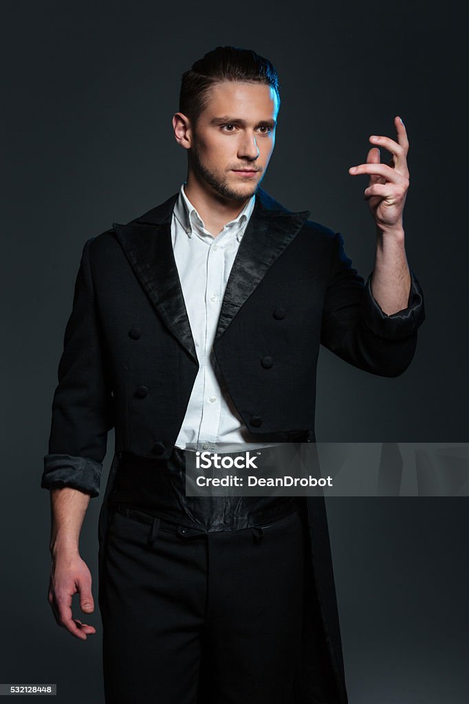 Serious young man magician in black tail coat showing tricks Serious young man magician in black tail coat standing with raised hand and showing tricks over grey background Tail Coat Stock Photo