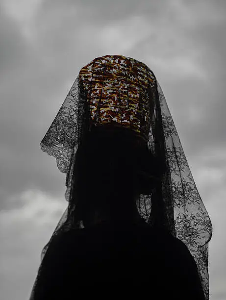 Woman wearing a headdress of black Spanish lace lifted high on a traditional comb standing with her back to the camera agaisnt a cloudy grey sky during a Holy Week in Andalucia, Spain