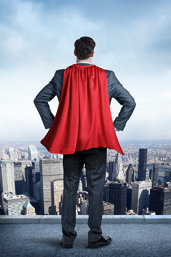 A businessman wearing a red cape looking out over the city below him. His hands are on his hips.  Blue sky and patchy clouds are in the distance.