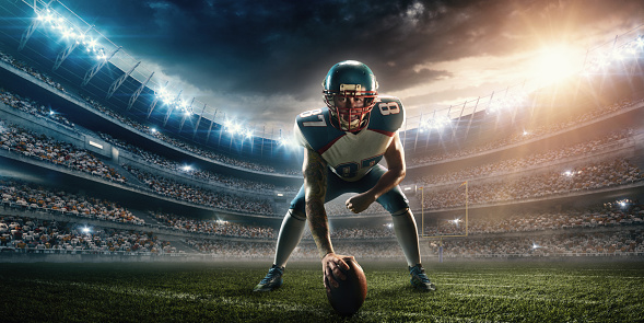 A male american football player in playing stand. He wears unbranded professional clothes. Action takes place in a generic outdoor football stadium under a cloudy sky with bright sun. 