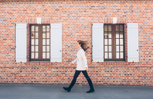 Smiling young woman walking on street. Wears white coat, black trousers and boots. Passing by old architecture building brick wall with opened vintage windows.