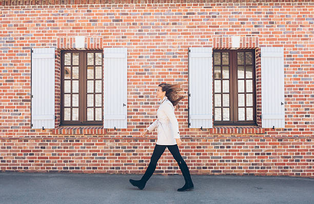 Portrait of young woman walking on street Smiling young woman walking on street. Wears white coat, black trousers and boots. Passing by old architecture building brick wall with opened vintage windows. wall sidewalk city walking stock pictures, royalty-free photos & images