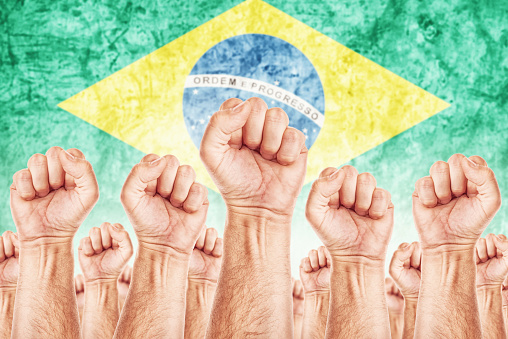 Brasil Labor movement, workers union strike concept with male fists raised in the air fighting for their rights, Brasilian national flag in out of focus background.