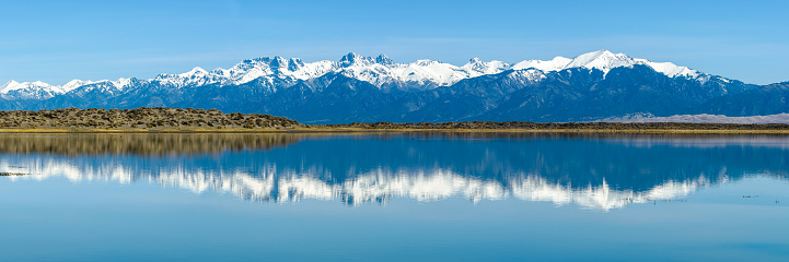 Panoramic view of Snow-capped Sangre de Cristo Mountains reflecting in San Luis Lake. The high peaks at the middle are: Challenger Point, Kit Carson Peak, Columbia Point, Crestone Peak, Crestone Needle, Music Mountain, Tijeras Peak, Cleveland Peak, left to right.