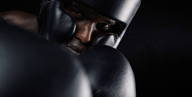 Close-up of african male boxer exercising Close-up image of african male boxer wearing protective head guard and gloves against black background. Young man exercising boxing. fist human hand punching power stock pictures, royalty-free photos & images