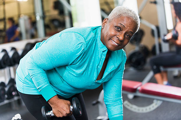 Senior African American woman at gym lifting weights Active senior staying fit.  Close up of African American woman (60s) at the gym, lifting dumbbell.  Bending over, looking at camera, smiling, happy expression on her face. exercise machine stock pictures, royalty-free photos & images