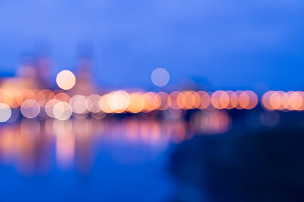 This image of the River Danube in Budapest, captures a blurred view of city lights at night, with warm orange and yellow bokeh orbs reflecting on a calm dark water surface, suggesting a river or harbor in an urban environment. The background features indistinct silhouettes of buildings and structures, creating an atmospheric and mysterious mood of nocturnal city life. No people are visible, emphasizing the abstract beauty of illuminated urban landscapes. Ideal for commercial uses such as backgrounds in articles on city development, nightlife scenes, travel promotions for metropolitan areas, or editorial content about modern architecture and evening ambiance in stock photography for media companies and authors.