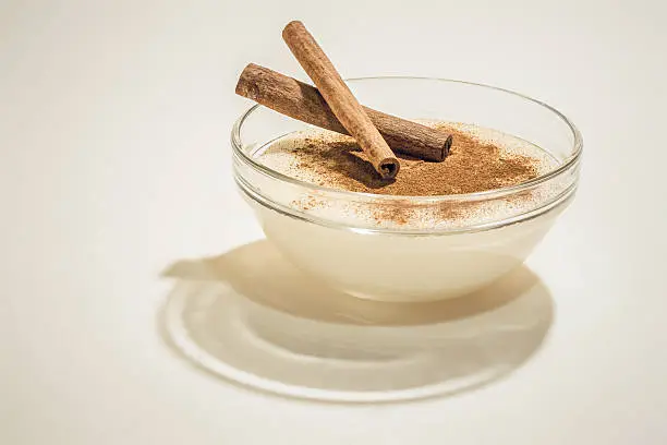 Creamy pudding in transparent glass bowl with cinnamon powder and cinnamon sticks on top, isolated on white background.
