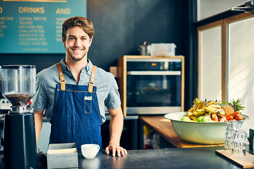 Portrait of confident barista standing at cafe counter. Young man is smiling in coffee shop. He is wearing denim apron.