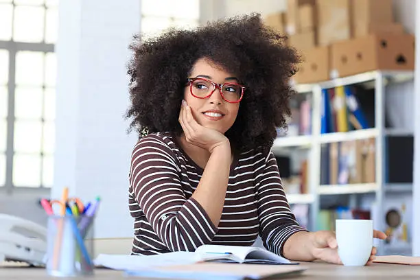 Smiling young woman with red eyeglasses drinking coffee at the office. Hand on chin and holding a coffee cup, looking away. As background shelves with boxes and folders, and tall windows. Office tools, note book and landline phone on desk.