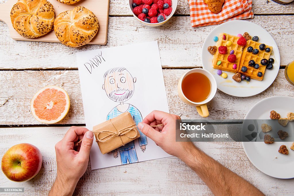 Childs drawing of her dad. Fathers day. Breakfast meal. Fathers day composition. Hands of unrecognizable man with his childs drawing of him, unpacking little present. Breakfast waffles with fruit. Studio shot on wooden background. Adult Stock Photo