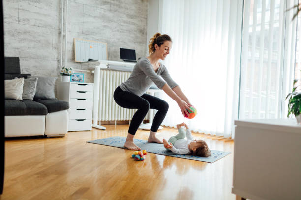Mother Exercise With Her Baby At Home Shot of an attractive young woman with her baby girl while doing exercise. Woman working squats and baby lying on exercise mat. They exercise in a living room. Mother playing with her baby while exercise, giving her baby ball. baby squatting stock pictures, royalty-free photos & images