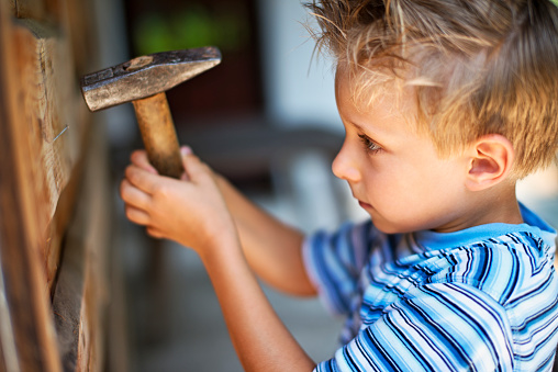 Little boy aged 6 learning to hammer a nail to the wooden cabin wall.