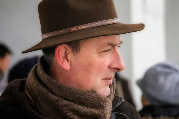 Middle age man with brown hat with rim and scarf during cold winter day looks into the distance. Serious expression of his face. Photo taken during busy day in city.