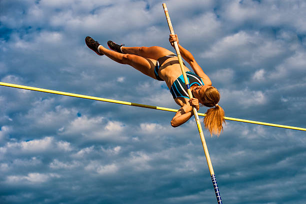 Young Women Jumping Over the Lath Against Cloudy Sky Young female athlete in pole vault action against the cloudy blue sky and late afternoon sunlight womens field event stock pictures, royalty-free photos & images