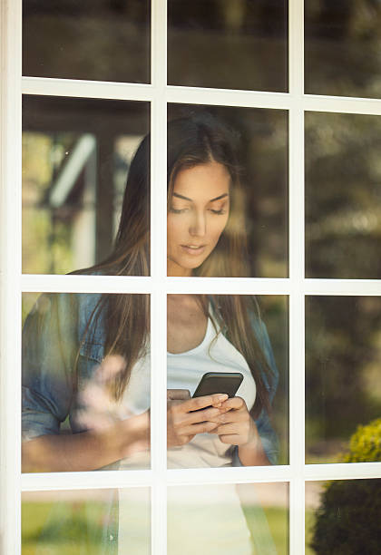 giovane donna in piedi dietro una finestra con telefono - mullion windows foto e immagini stock