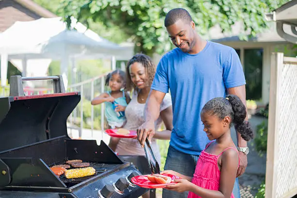 A family of four are outside on a beautiful sunny summer day. They father is barbecuing hot dogs and hamburgers over the grill.