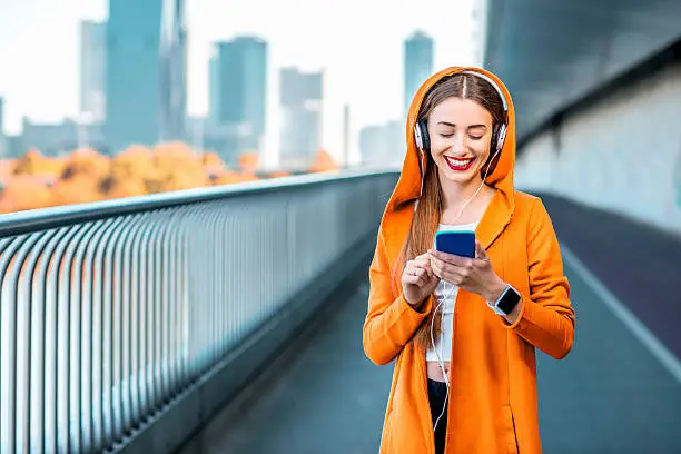 Young sport woman in yellow sweater listening to the music with smart phone on the modern bridge with skyscrapers on the background. Morning exercise in megacity
