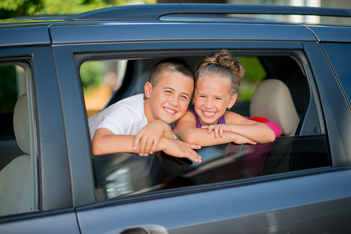 A brother and sister are sitting in their parents van and are looking out the window. They are both smiling and looking at the camera.