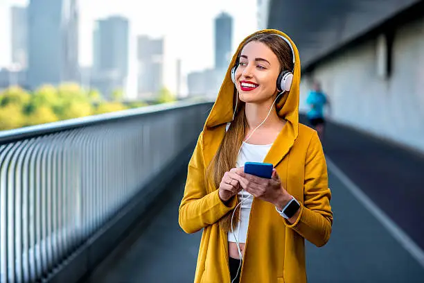 Young sport woman in yellow sweater listening to the music with smart phone on the modern bridge with skyscrapers on the background. Morning exercise in megacity