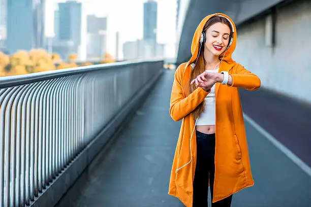 Young sport woman in yellow sweater listening to the music with smart watch on the modern bridge with skyscrapers on the background. Morning exercise in megacity