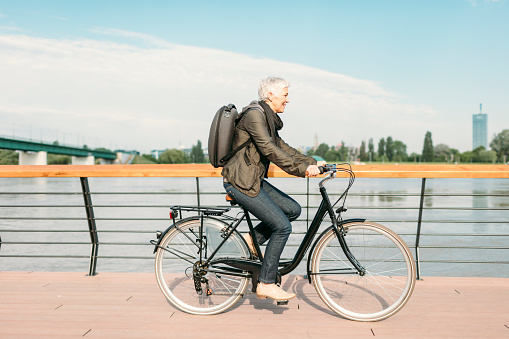 Mature businesswoman riding bicycle to and from her office. She is riding near river. Casual clothing with modern business backpack on her back. Riding city style bike.