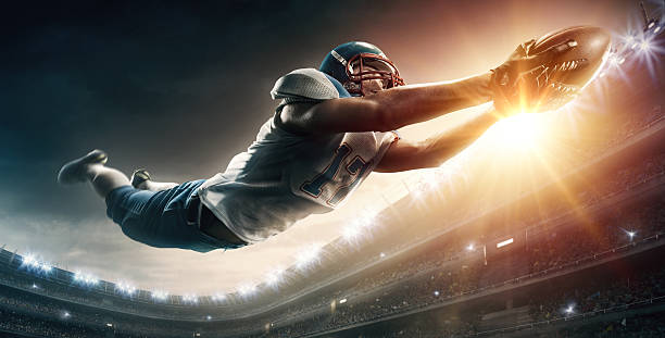 American football player jumping Wide angle low view of an American Football player jumping with a ball in the end zone of a Football field for a touchdown. The action takes place on professional stadium. The player wears unbranded sports uniform. football helmet and ball stock pictures, royalty-free photos & images