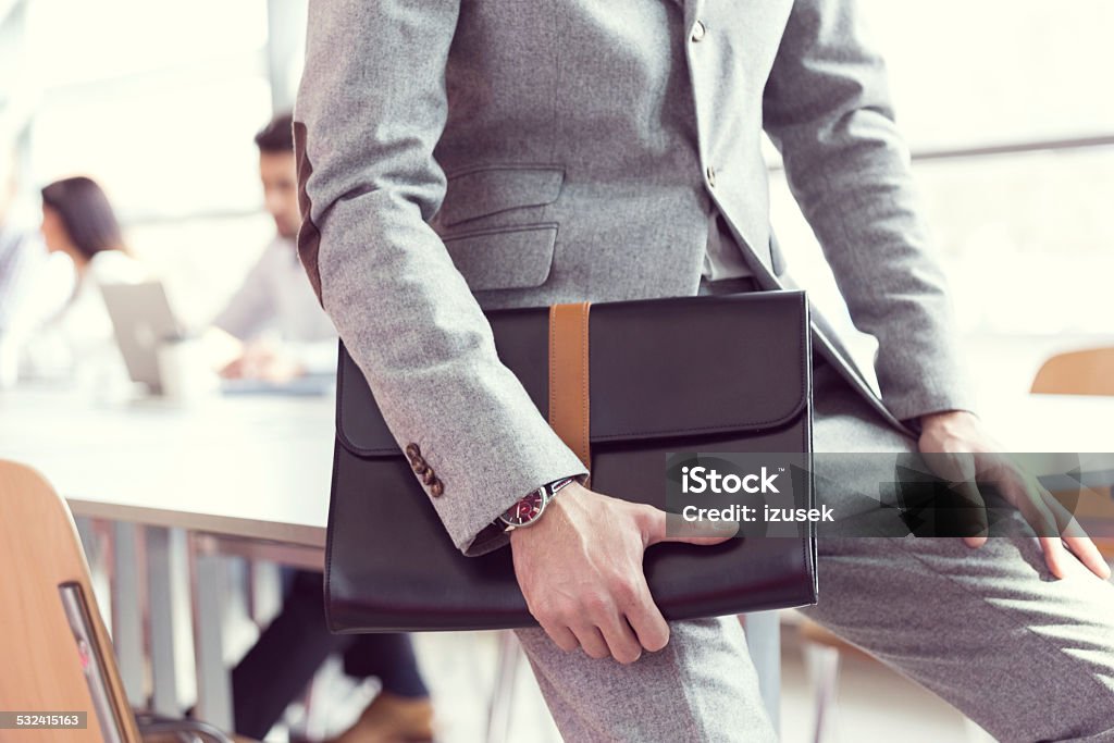 Businessman wearing suit holding briefcase Close up of businessman sitting in a meeting room, wearing grey suit, holding leather briefcase with business people working in the background. 2015 Stock Photo