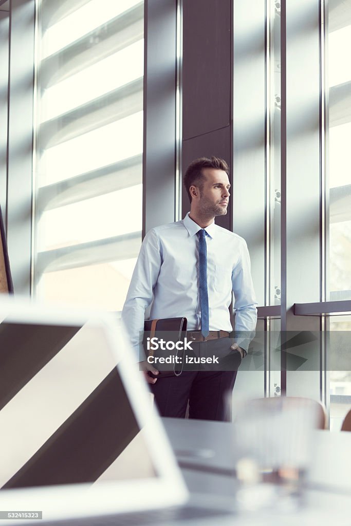 Confident businessman in office, holding a briefcase Portrait of confident businessman wearing shirt and tie standing by the window in an office, holding a leather briefcase in hand. Business Person Stock Photo