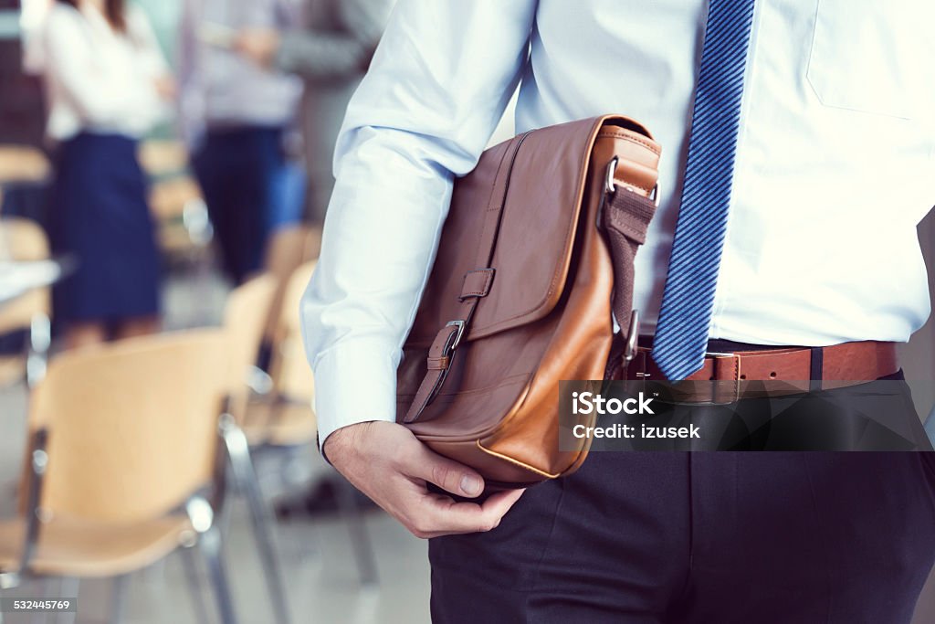 Businessman holding briefcase, close up Businessman standing in a meeting room, wearing shirt and tie, holding leather briefcase with coworkers in the background. Close up of hands. Briefcase Stock Photo