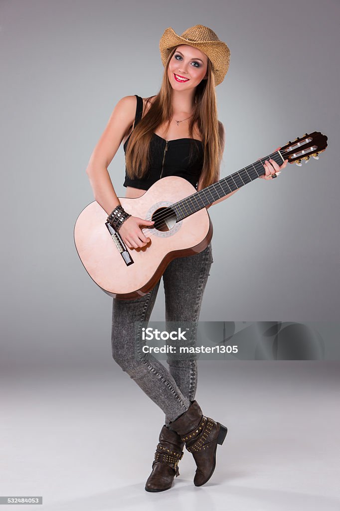 The beautiful girl in a cowboy's hat and acoustic The beautiful girl in a cowboy's hat playing acoustic guitar on a gray background. Portrait in full growth Acoustic Guitar Stock Photo