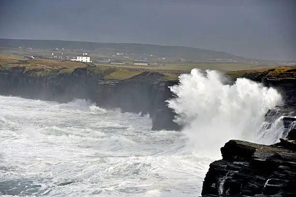 Big seas pound the huge black cliffs near Doolin, from waves that surge across the wild Atlantic Ocean, dwarfing an Irish mansion perched on the grassy cliffs and glowing in the setting sun. The power of the rising seas is echoed in the frothy spume that coats the water's surface.