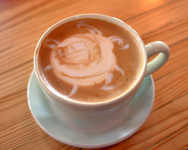 A hot espresso latte in a modern ceramic mug with a turtle poured into the steamed milk foam. View from directly above on a wood grain table background.