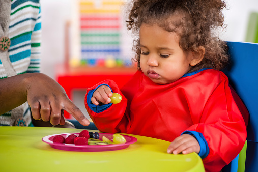 A portrait of an African American carer engaging a toddler to eat other fruit.