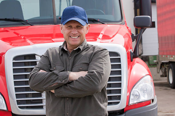 A royalty free image from the trucking industry of a smiling and friendly truck driver standing in front of his red tractor trailer at a warehouse.
