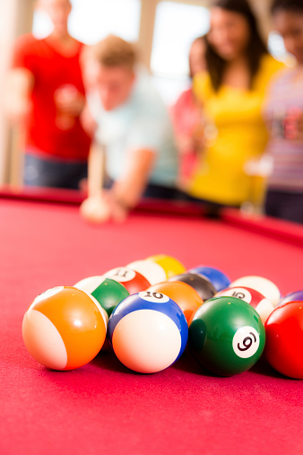 Focus on unbroken billiards balls on red felt pool table. Young adult, multi-ethnic group of friends are in the background. Young man is about to break the balls with cue ball, stick. Colorful.  