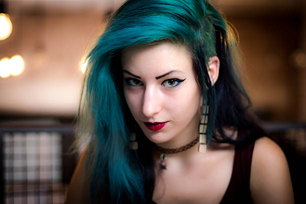 A close-up portrait shot of a teenage girl with short green-dyed hair standing outdoors, looking to the side with a serious expression, wearing a casual white t-shirt while leaning against a modern green wall background.Videos are available similar to this scenario.