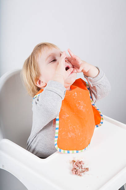 shouting in lunch blonde caucasian baby seventeen month age orange bib grey sweater eating tuna rice on white high-chair shouting with hands kids eating tuna stock pictures, royalty-free photos & images