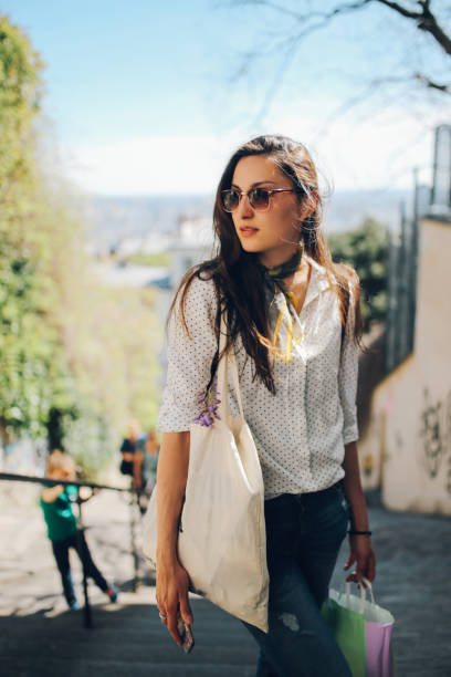 Vintage toned image of a young Parisian woman walking home after work or shopping on the streets of Montmartre district of Paris. She is wearing a white dotted pattern shirt and sunglasses, carrying a canvas tote bag and some shopping bags on a bright and sunny day. Paris, lifestyle, street style fashion concepts.