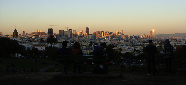 San Francisco, California, USA - November 24th, 2013: a group of visitors is sitting on a bench whereas other two are standing on a city park hill while thay are watching the sunset reflecting on the downtown skyline on a clear sky Autumn evening, in Mission Dolores District, San Francisco
