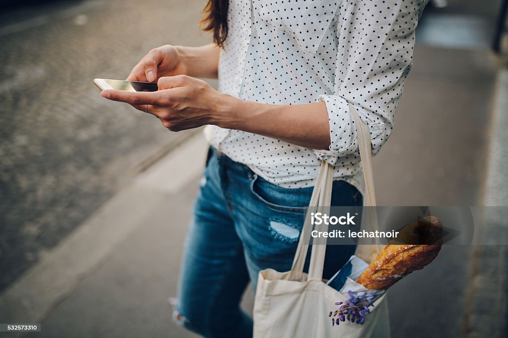 Young Parisian woman using the smartphone Vintage toned image of a young Parisian woman using her smartphone while walking the streets on Montmartre district of Paris. She is wearing a white dotted pattern shirt and sunglasses, carrying a canvas tote bag after some shopping for groceries and food. Paris, lifestyle, street style fashion concepts. Supermarket Stock Photo