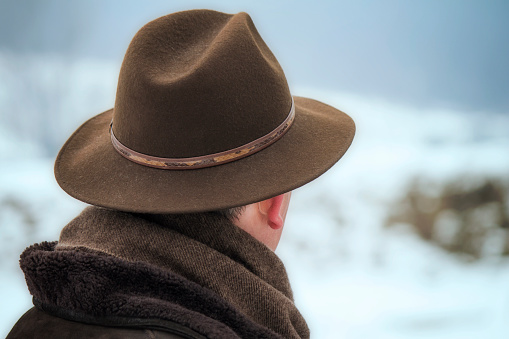 Well-dressed solitary man with brown hat looks into the distance.  Winter ambient during day in nature with gray sky over snowy hill.