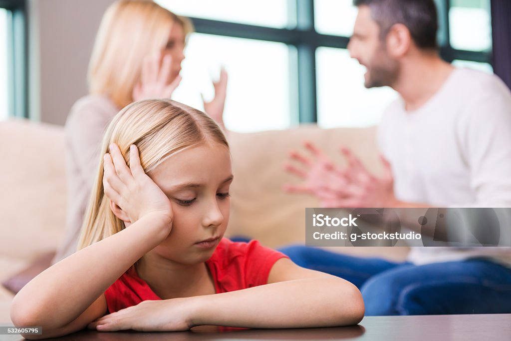 I hope they will work it out. Sad little girl leaning at the table and holding head in hand while her parents shouting at each other in the background Girls Stock Photo