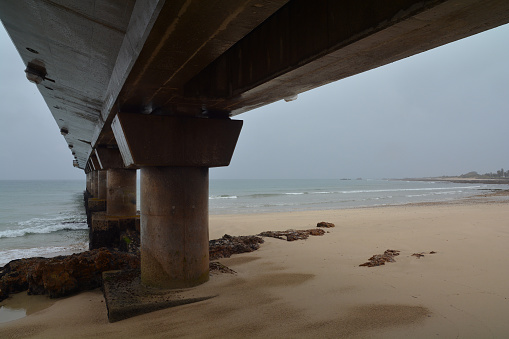 The under belly of a large concrete structure seaside pier on a very rainy day with a deserted beach.