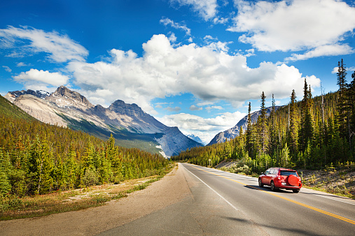 Tourist red car taking a road trip, driving through Canadian Rockies, Banff National Park, Alberta, Canada. Summer vacation travelers seek North American scenic natural destinations to experience mountain range landscape views, snow-capped glacial peaks, majestic, tranquil wilderness areas, and beauty in nature. Sunny highway and cloudy sky provide copy space.