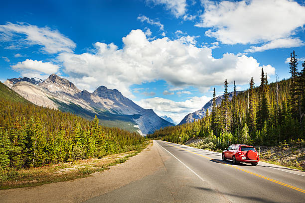 Banff National Park Road Trip Drive through Canadian Rocky Mountains Tourist red car taking a road trip, driving through Canadian Rockies, Banff National Park, Alberta, Canada. Summer vacation travelers seek North American scenic natural destinations to experience mountain range landscape views, snow-capped glacial peaks, majestic, tranquil wilderness areas, and beauty in nature. Sunny highway and cloudy sky provide copy space. canadian rockies stock pictures, royalty-free photos & images