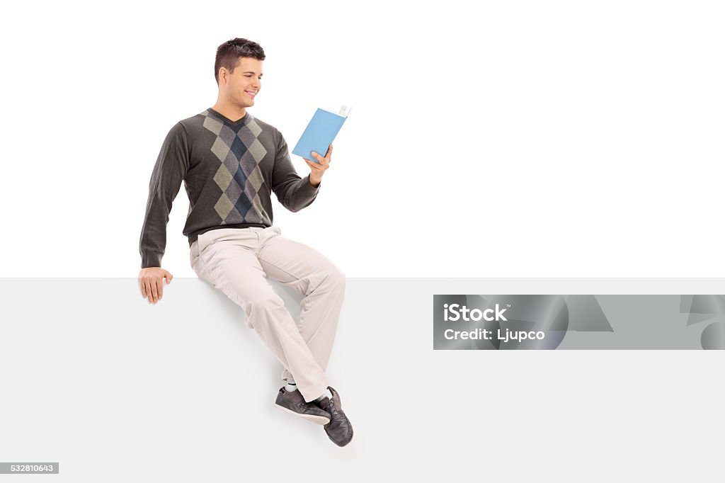 Casual guy reading a book seated on a blank panel Casual guy reading a book seated on a blank panel isolated on white background 20-29 Years Stock Photo