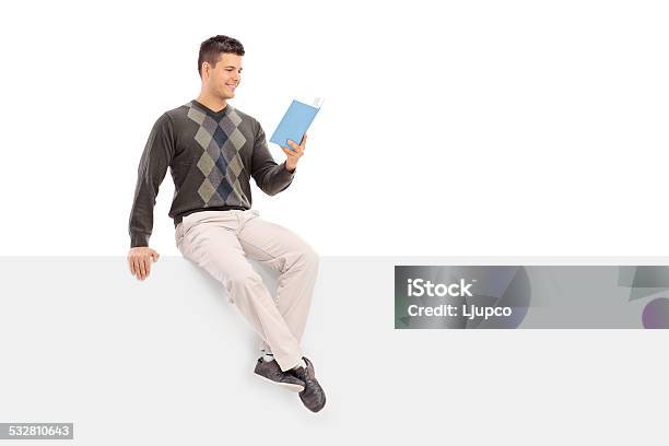 Casual Guy Reading A Book Seated On A Blank Panel Stock Photo - Download Image Now - 20-29 Years, 2015, Adult