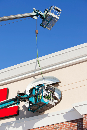 Two workers in an aerial platform are installing a letter S for a commercial building exterior sign. The sign is illuminated with LEDs which are more energy efficient than conventional lighting and are becoming commonplace with new building projects.